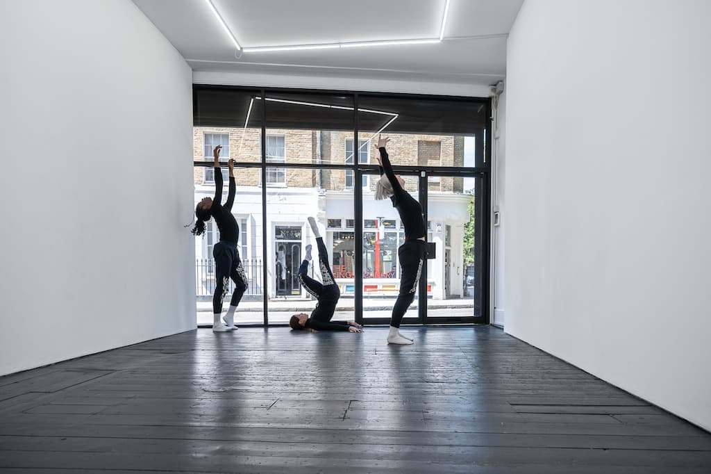 Three performers, dressed in black, two stading, one upside down on the floor, enacting A Cricle Whispering Dot by Nicole Bachmann