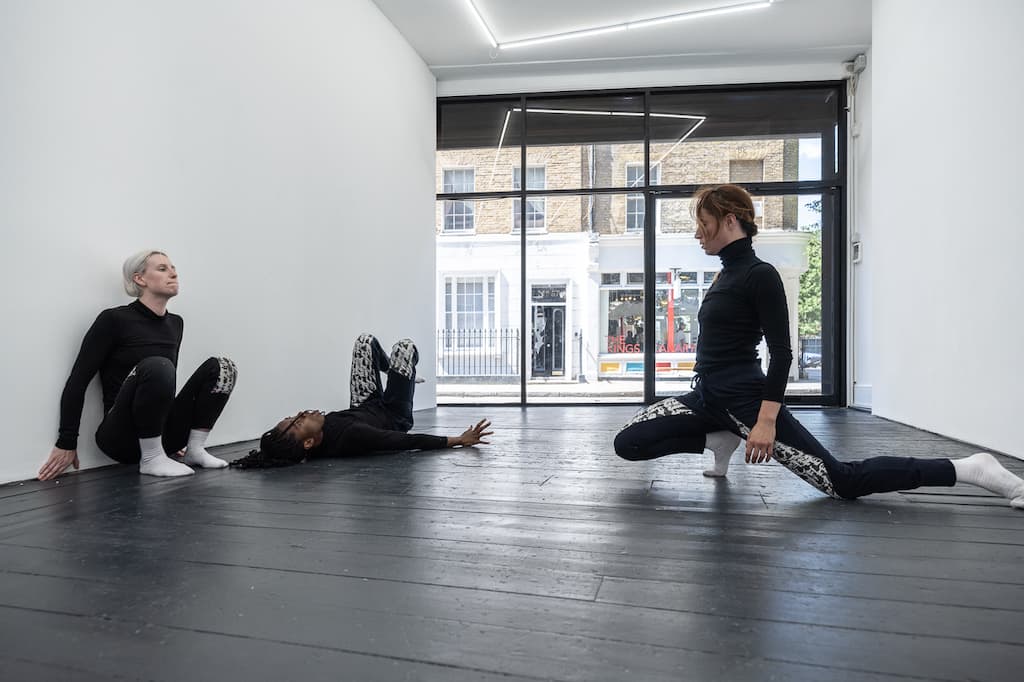 Three performers, dressed in black, one on the floor, one kneeling, the other sqatted against a wall, enacting A Cricle Whispering Dot by Nicole Bachmann