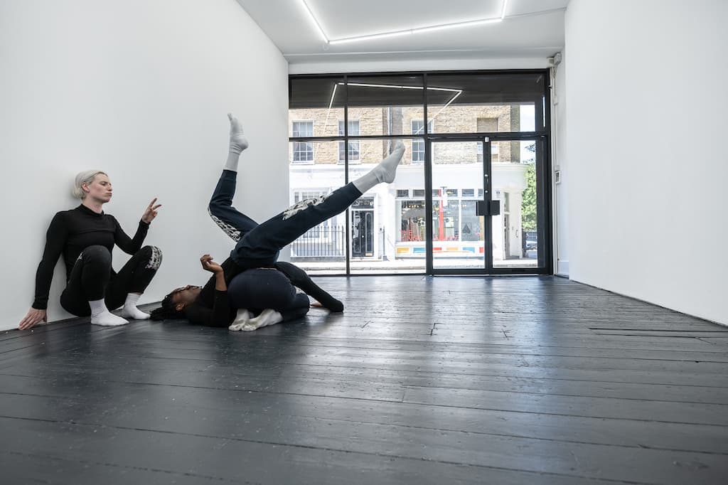Three performers, dressed in black, one crouched on the floor, another doing a upside down on top of them, the third sqatted against a wall, enacting A Cricle Whispering Dot by Nicole Bachmann