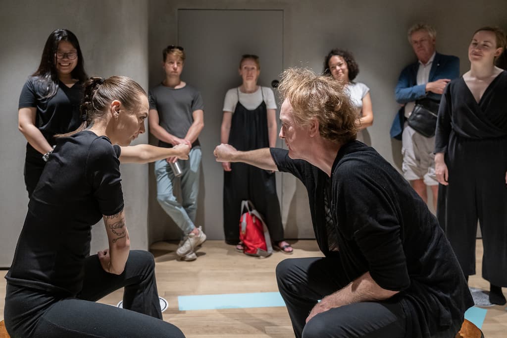 Installation shot showing two performers sat in the middle of the pentagram with one arma out to the side mirroring each others pose. Audience is wathcing in the background.