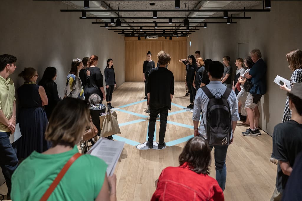 Performers, dressed in black, stood around a pentagram shape painted on the floor. Audience is watching in the foreground.