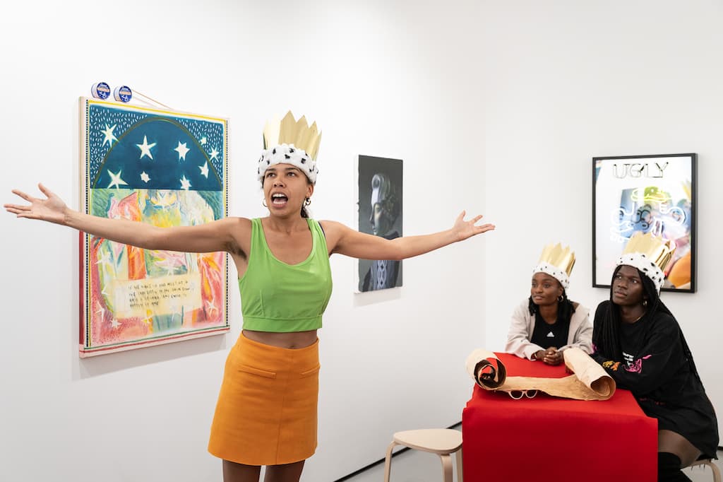 Female performer with arms stretched being wathched by two others sat at a table.