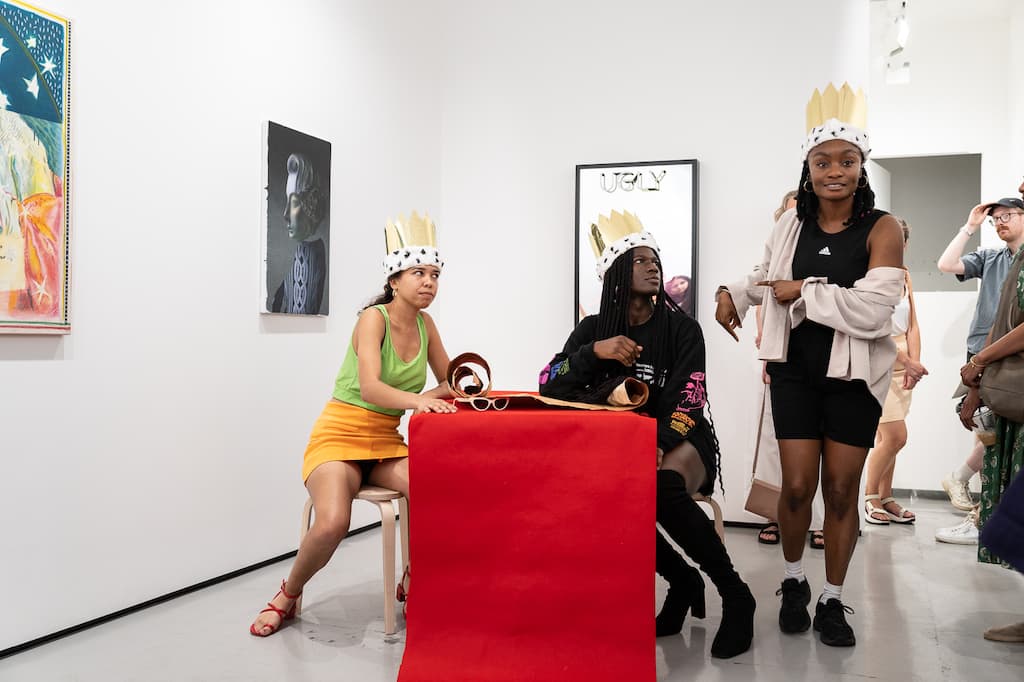 Three performers, all wearing crowns grouped around a table. Two are sitting.