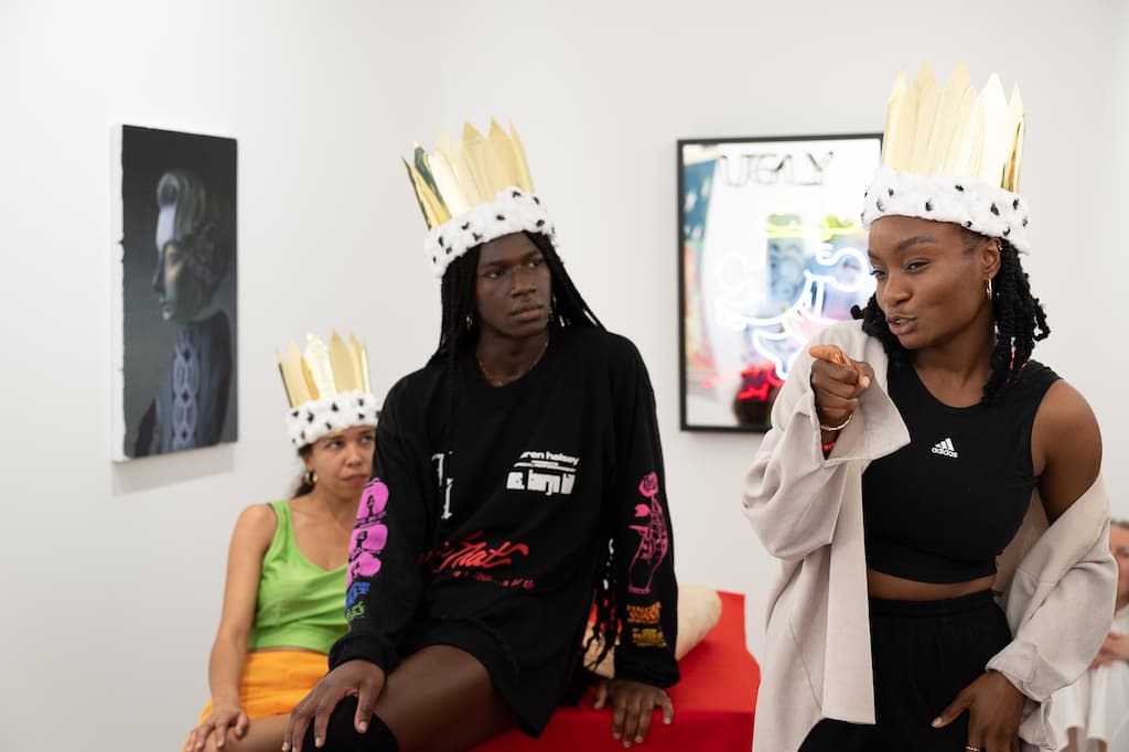 Three performers, all wearing crowns. One is sitting on a chair, the other on a table, the third is standing.