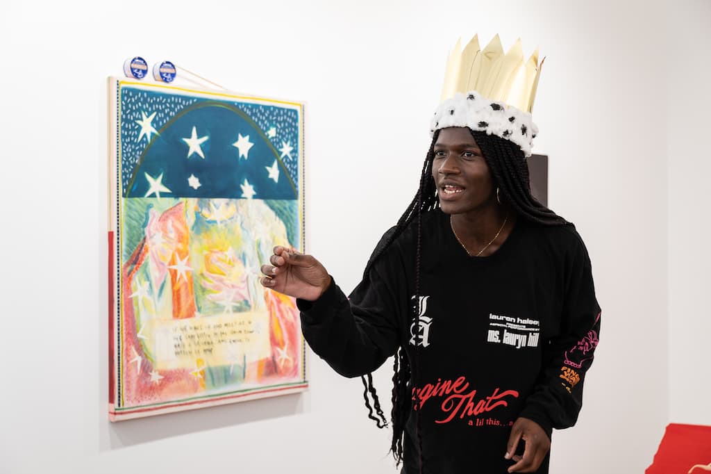 Performer, wearing a crown standing and gesticuling in front of a painting.